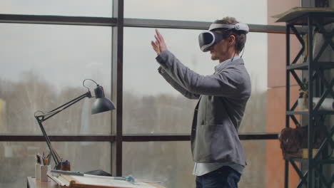 male-engineer-in-a-virtual-reality-helmet-near-the-large-window-with-his-hands-imitates-the-work-of-the-interface.-Designing-the-future-the-concept-of-virtual-architecture-and-design-interface-graphic-applications.